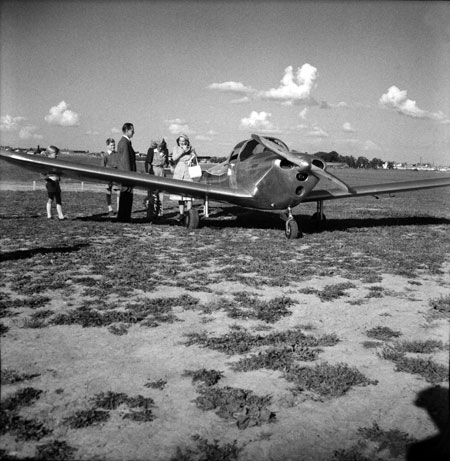 Ercoupe SE-AWU glittrar i solen. Bakom damerna skymtar en Cub. Bredvid nosen syns gamla Ekängs gård i en dunge. Pappersbrukets skorsten anas längre t.h. Foto: Åke Ahlstrand, Specialfoto