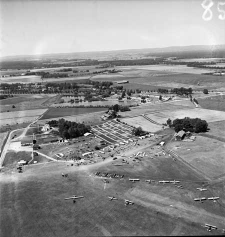 Gustavsvik 3 juli 1950 under VM i segelflyg. Massor av plan gästar. Bergslagens flygs hangar med vitt tak och Aero-Frabhs långa barack syns t.v. om de rutiga kolonilotterna. Gustavsviksbadet i bakgrunden. Foto: Åke Ahlstrand, Specialfoto